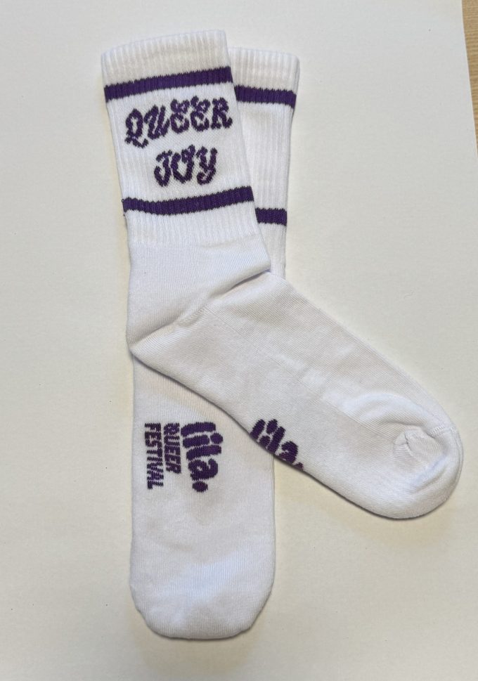 Weisse Socken mit eingestricktem Schriftzug «Queer Joy» oben und dem Logo «lia. Queer Festival» auf der Fussunterseite